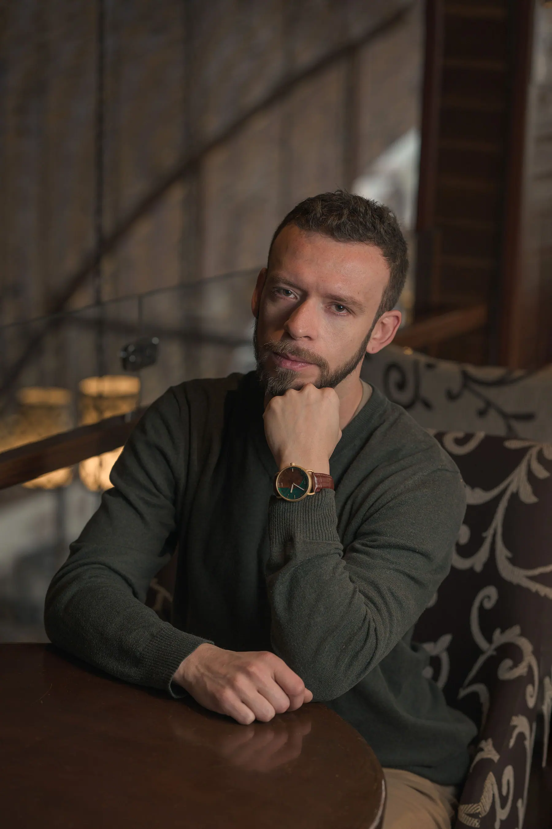 Leaning on the wooden bar of a café in an olive knit, hand at his chin, in warm interior light.