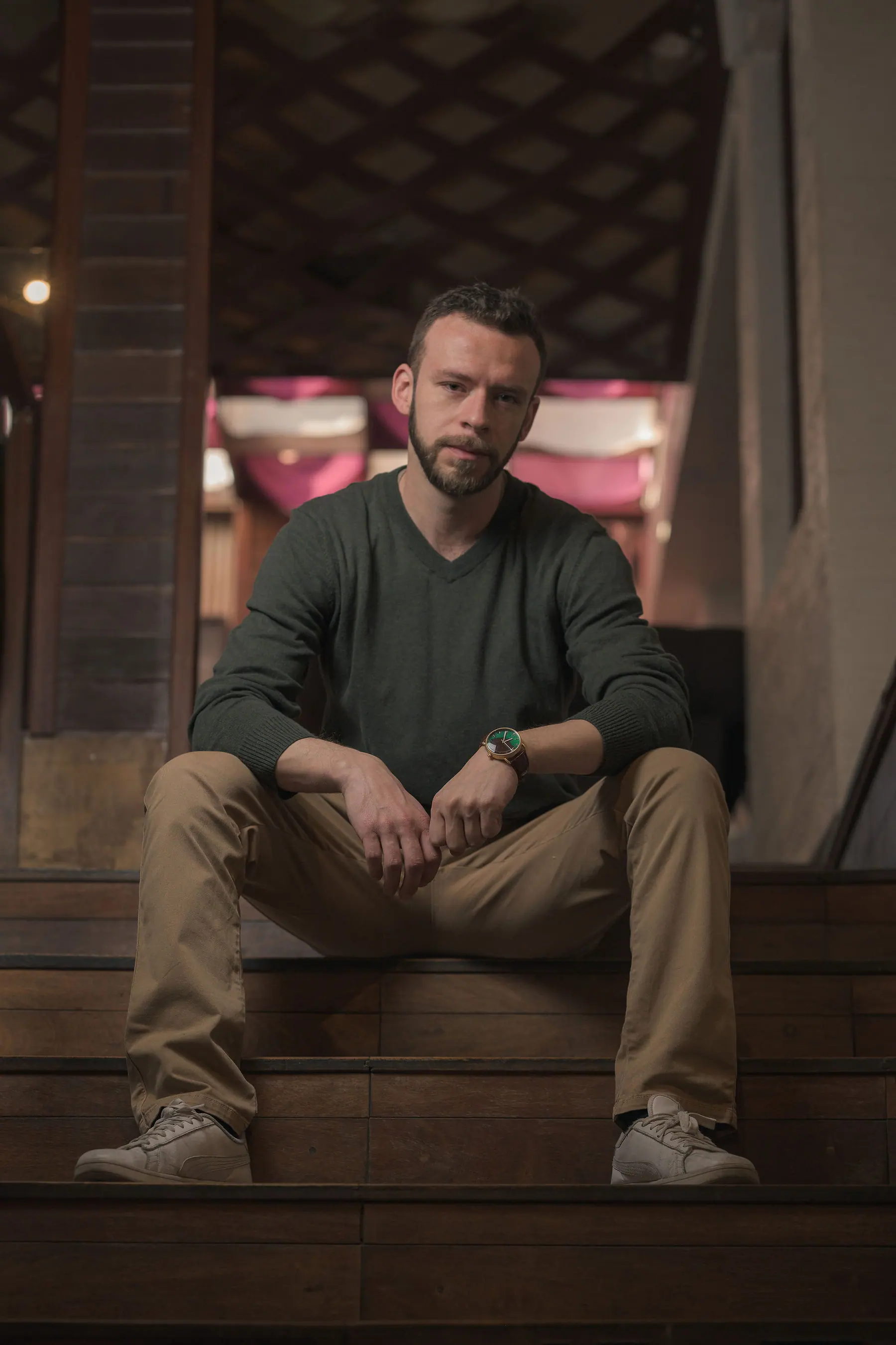 Sentado en unos escalones de madera con jersey verde oliva y chinos beige, en un interior cálido.
