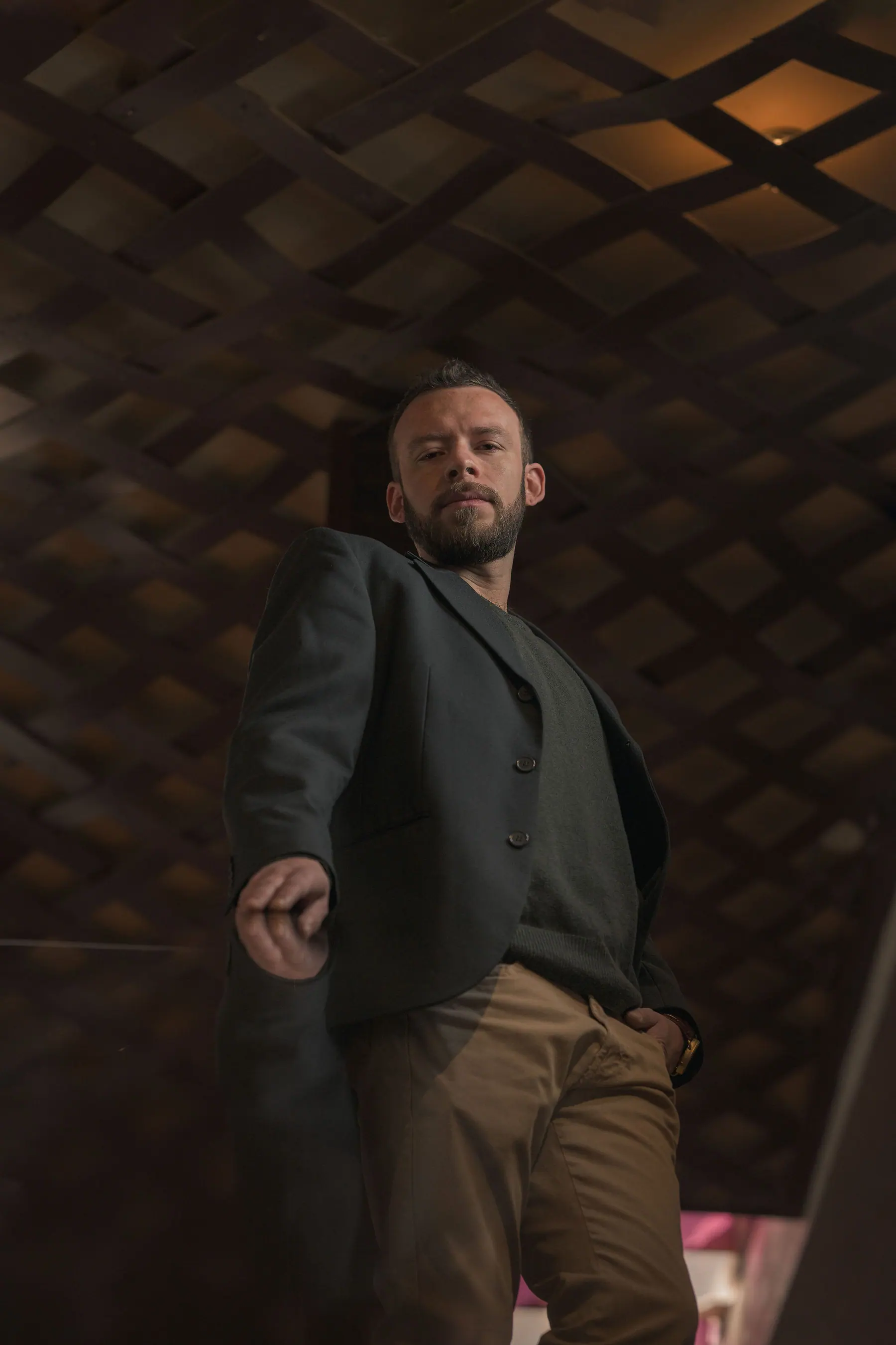 Elex Asbun walking toward the camera in a dark green blazer and camel trousers, before a dim wooden lattice.