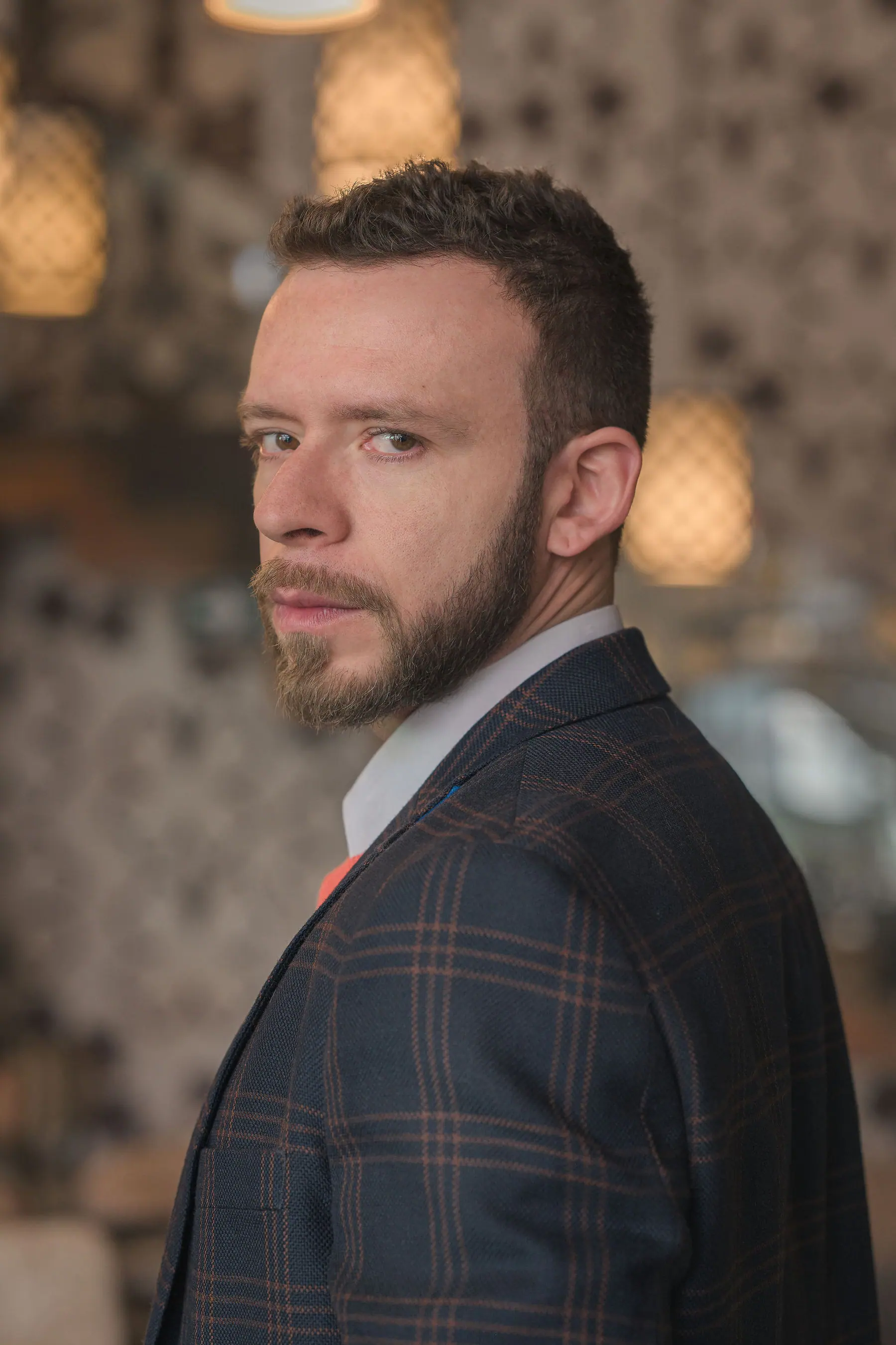 Retrato de medio cuerpo con blazer de cuadros azul, camisa blanca y jersey coral, con luces desenfocadas de un interior detrás.