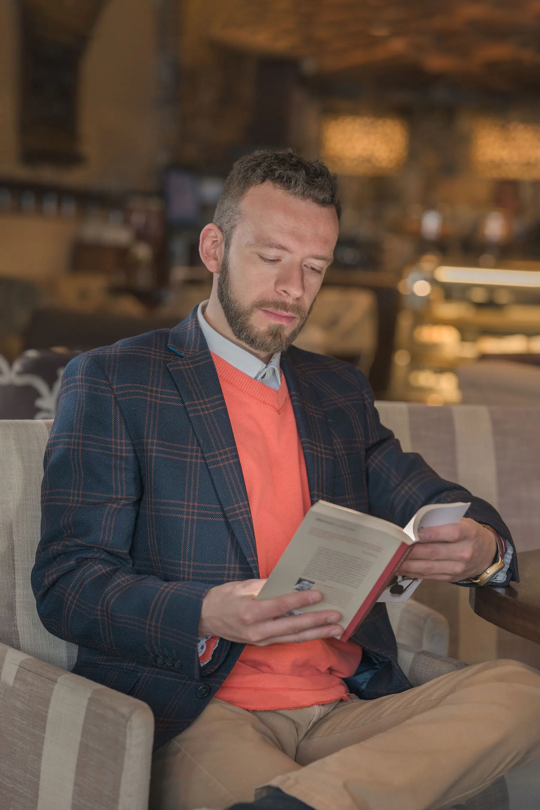 Elex Asbun reading a book in a café armchair, wearing a blue checked blazer and coral knit.