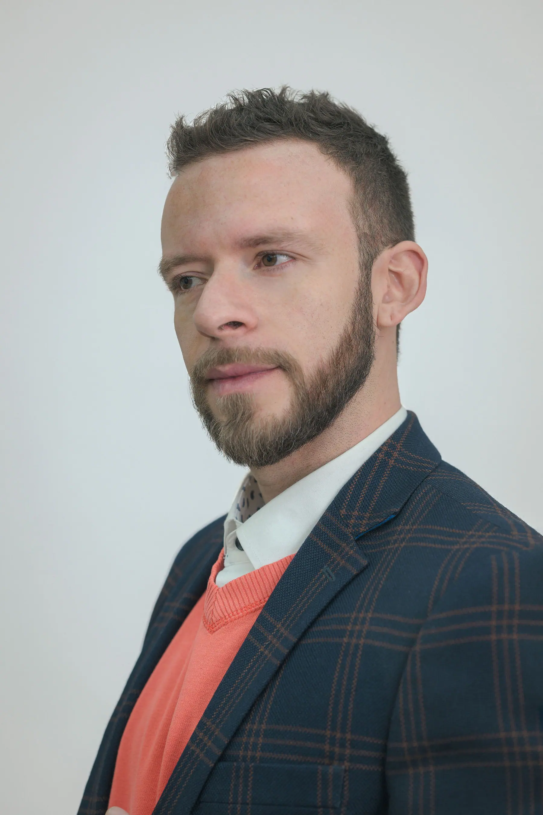 Retrato de estudio sobre fondo gris claro con blazer azul marino, camisa blanca y jersey coral.