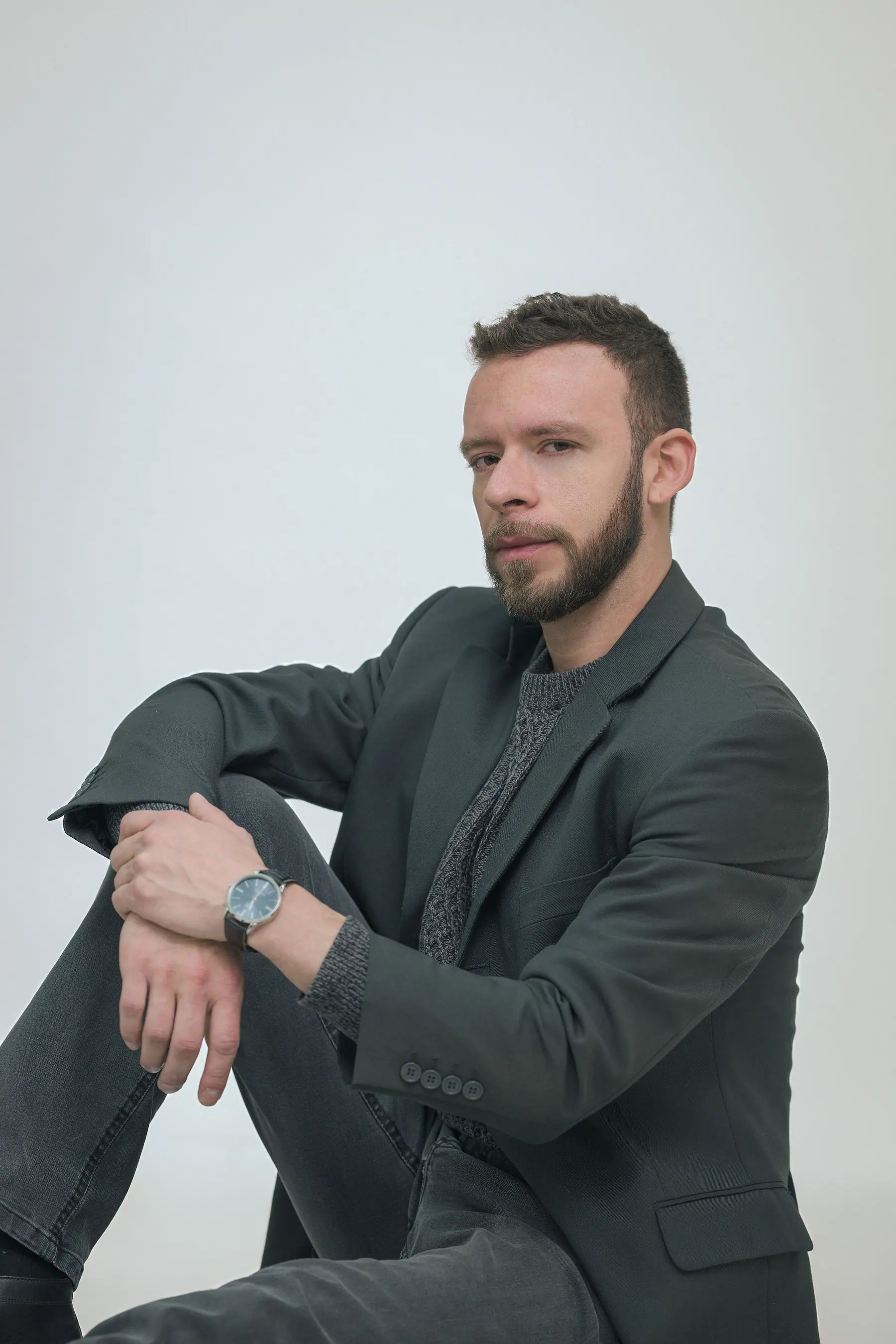 Sentado con el antebrazo sobre la rodilla y el reloj a la vista, blazer gris oscuro sobre fondo neutro de estudio.
