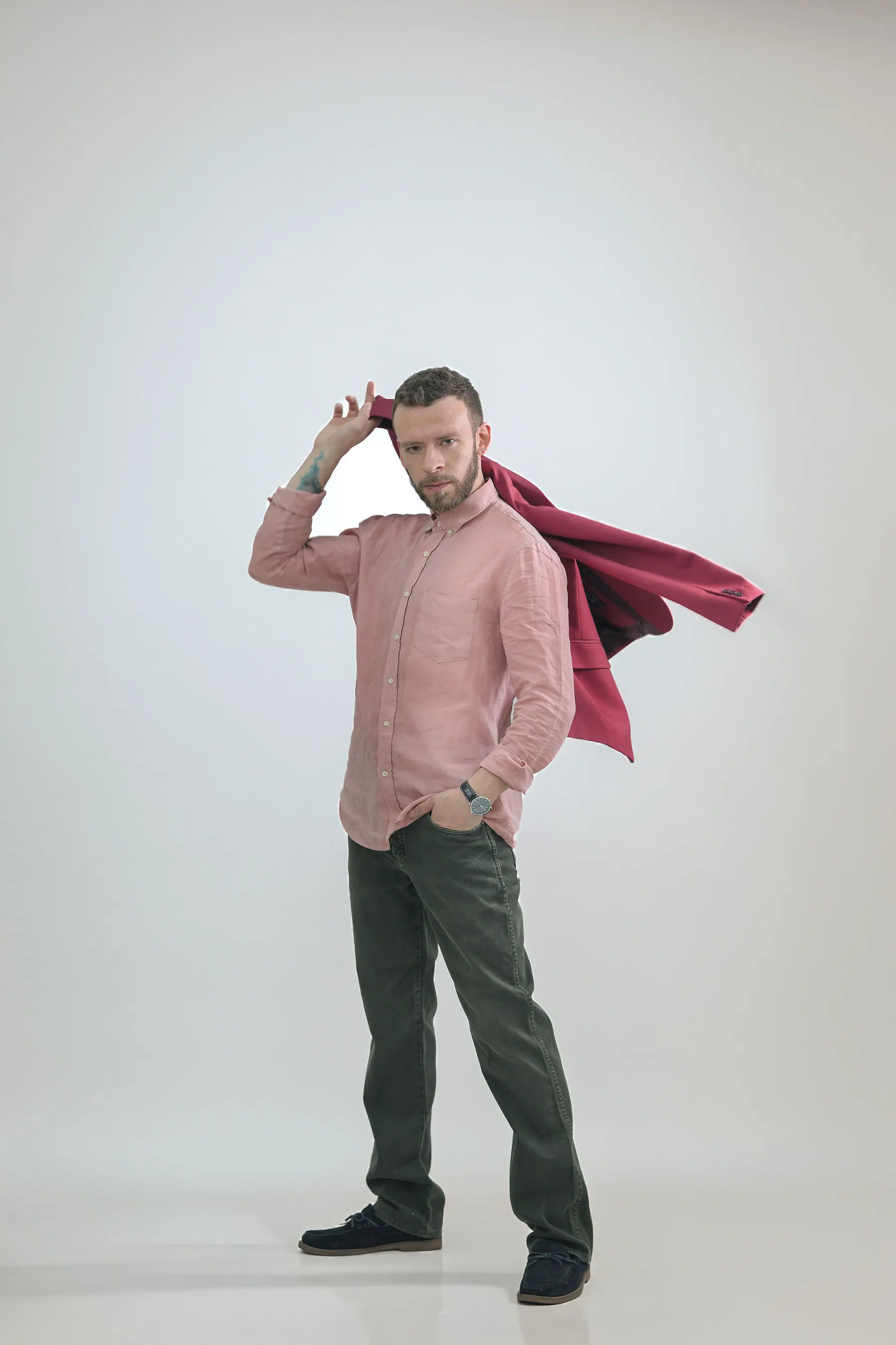 En estudio sobre fondo blanco, camisa rosa y pantalón verde oliva, con una chaqueta granate en el aire.