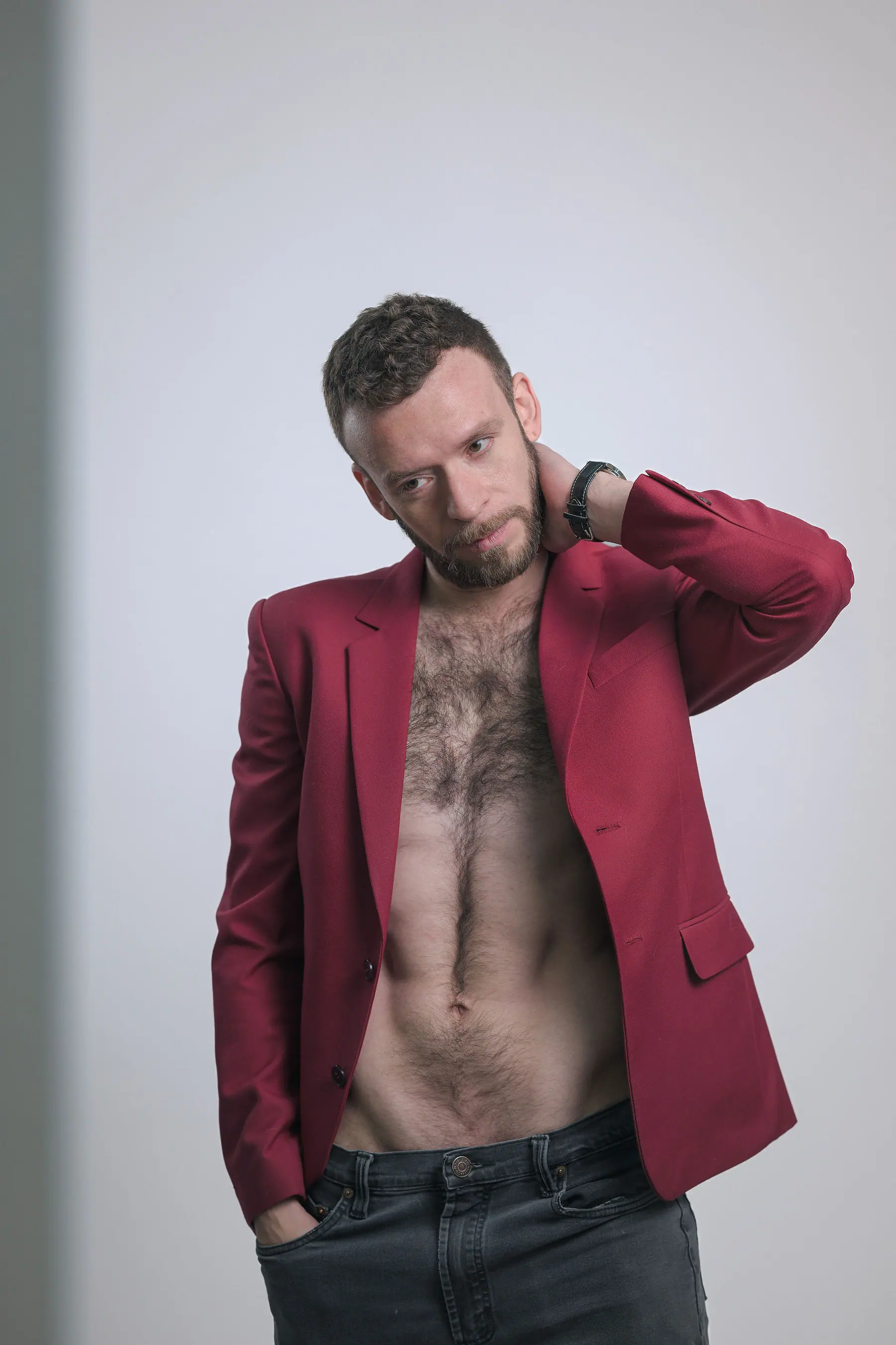 Elex Asbun in the studio wearing an open red blazer over a bare chest and dark jeans, hand at the back of his neck.