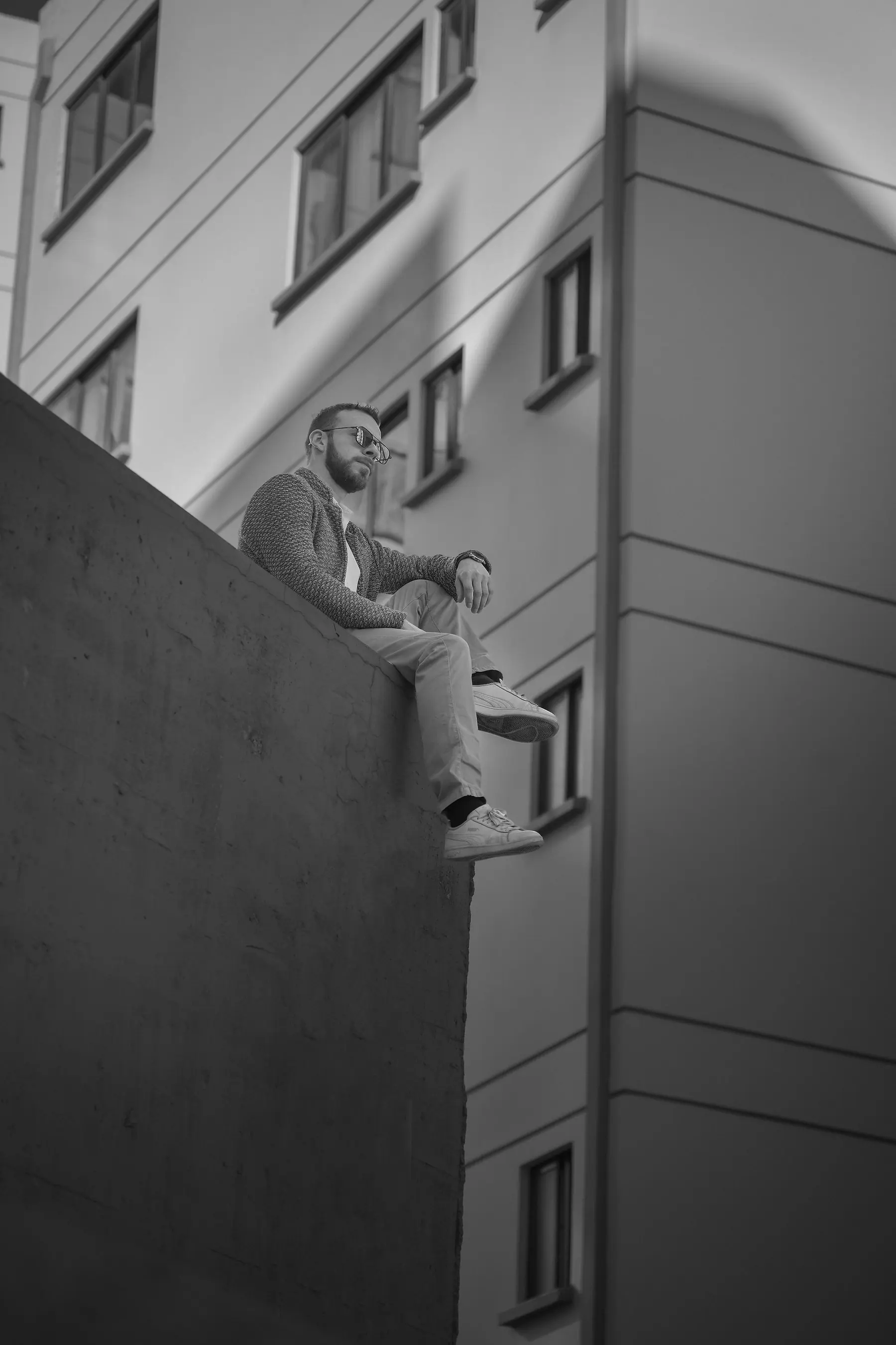 Black and white photograph of Elex Asbun seated on the edge of a high wall, a modern building facade behind him.