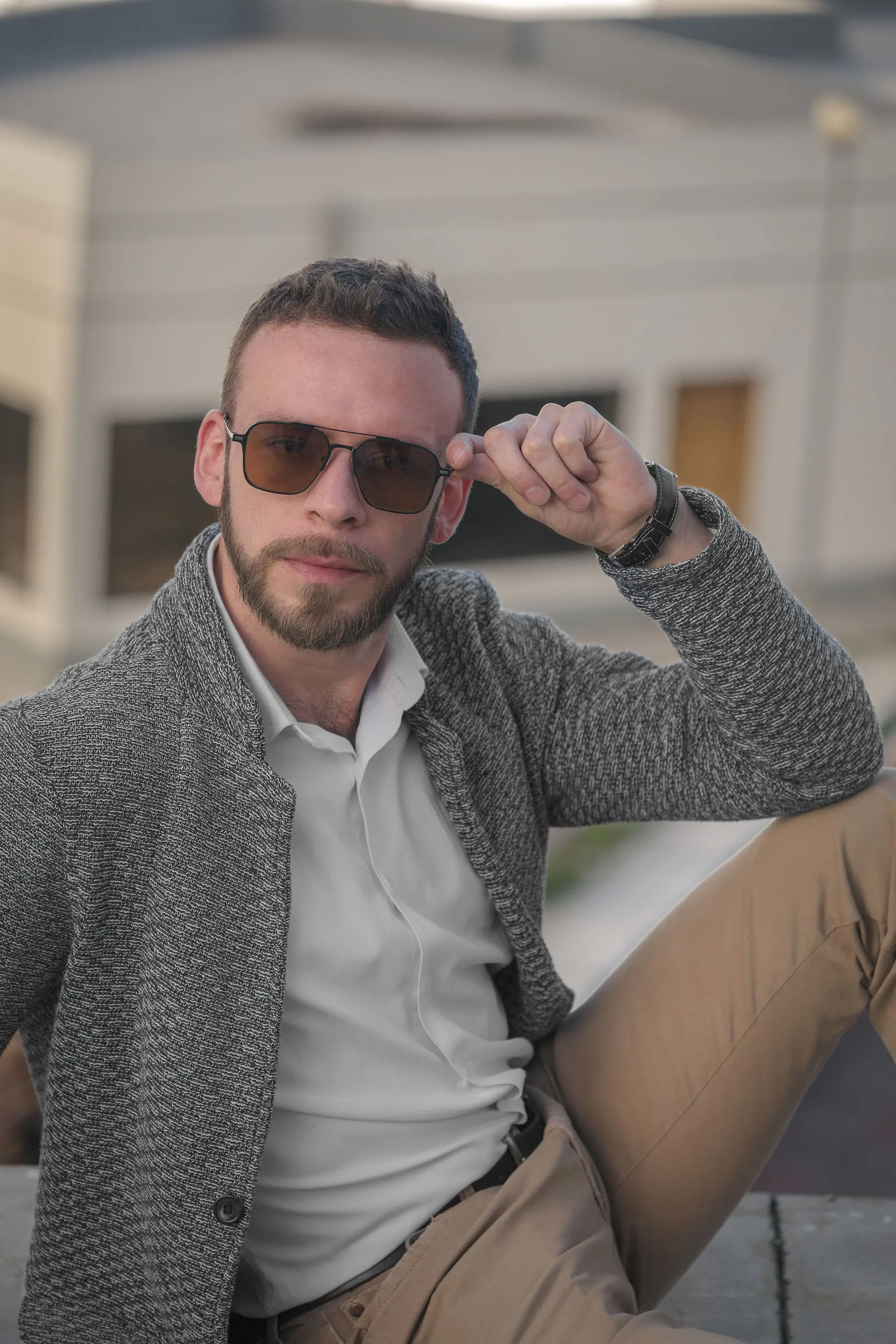 Sentado en exterior con gafas de sol, cárdigan gris de punto grueso, camisa blanca y chinos camel.