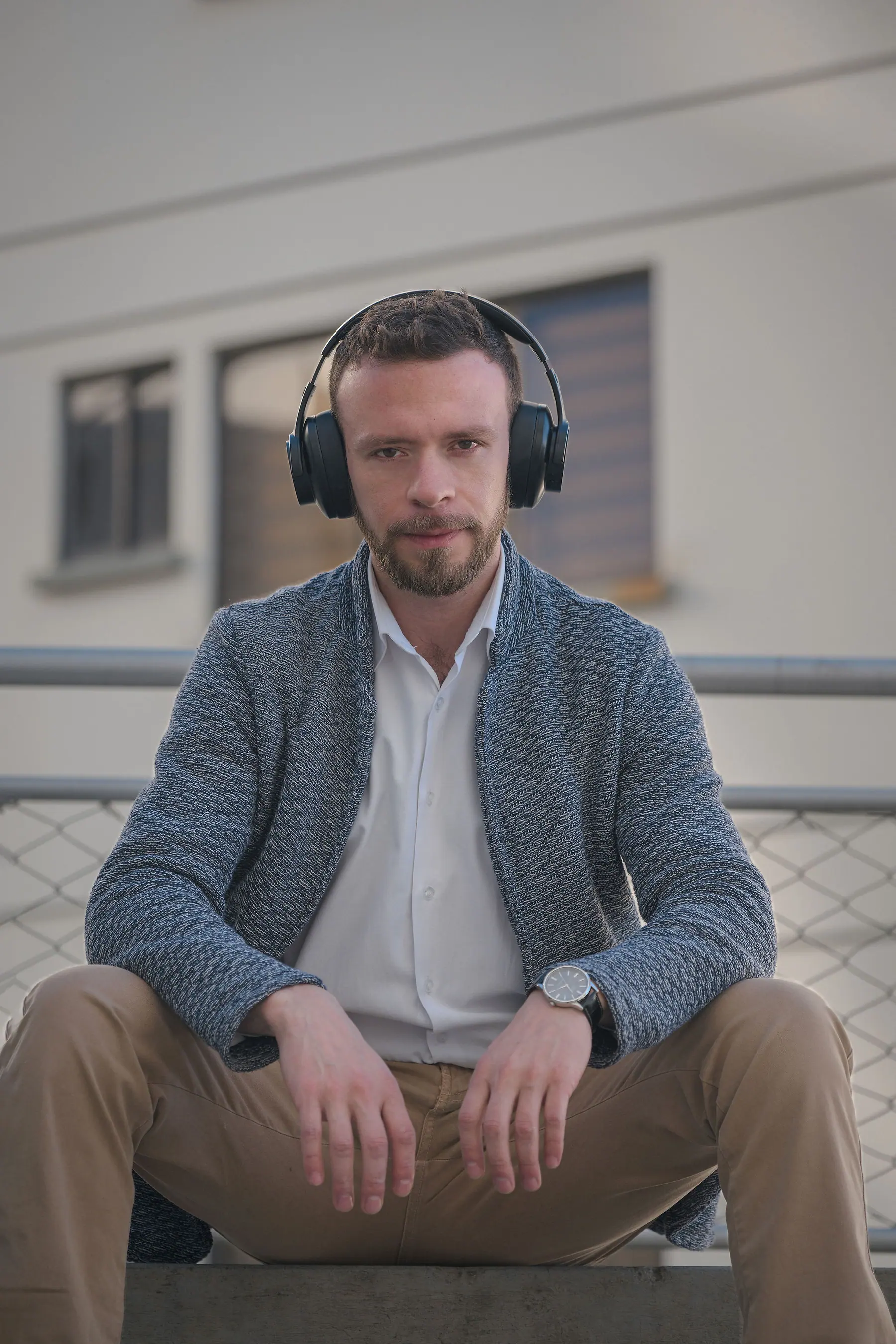 Seated outdoors wearing over-ear headphones, a blue knit cardigan, white shirt and camel chinos.