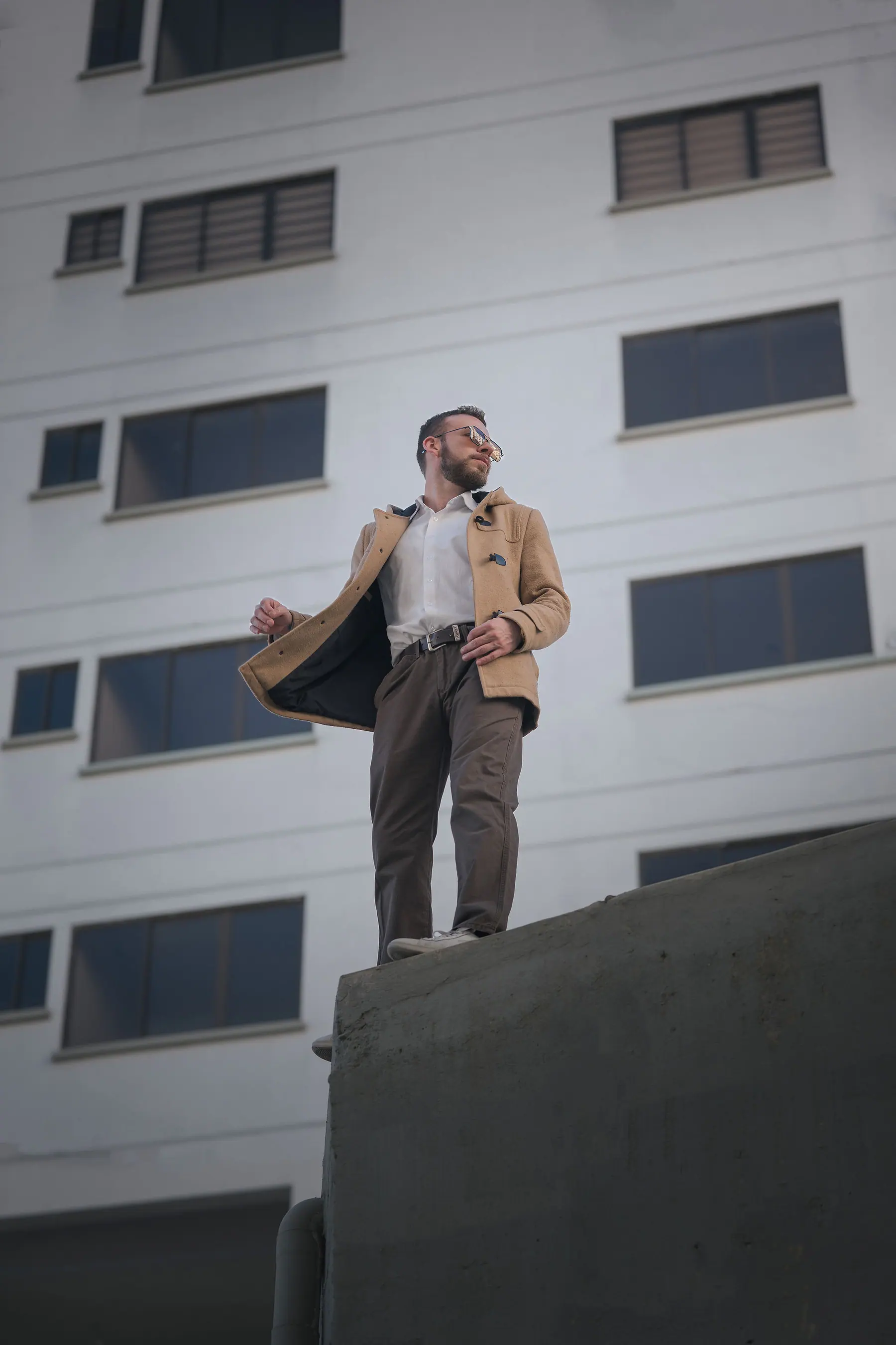 Standing on a wall in a camel coat open to the wind, before the white facade of a building with aligned windows.