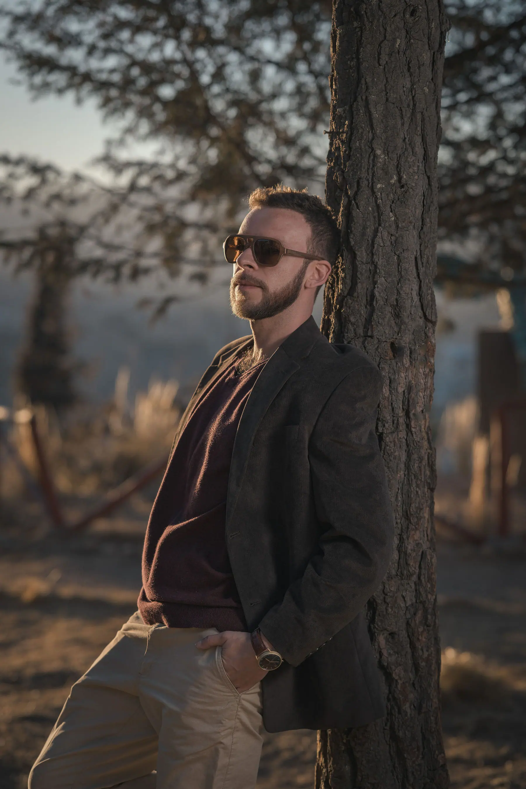 Elex Asbun in sunglasses, a brown blazer and burgundy knit, leaning against a pine trunk backlit by the setting sun.
