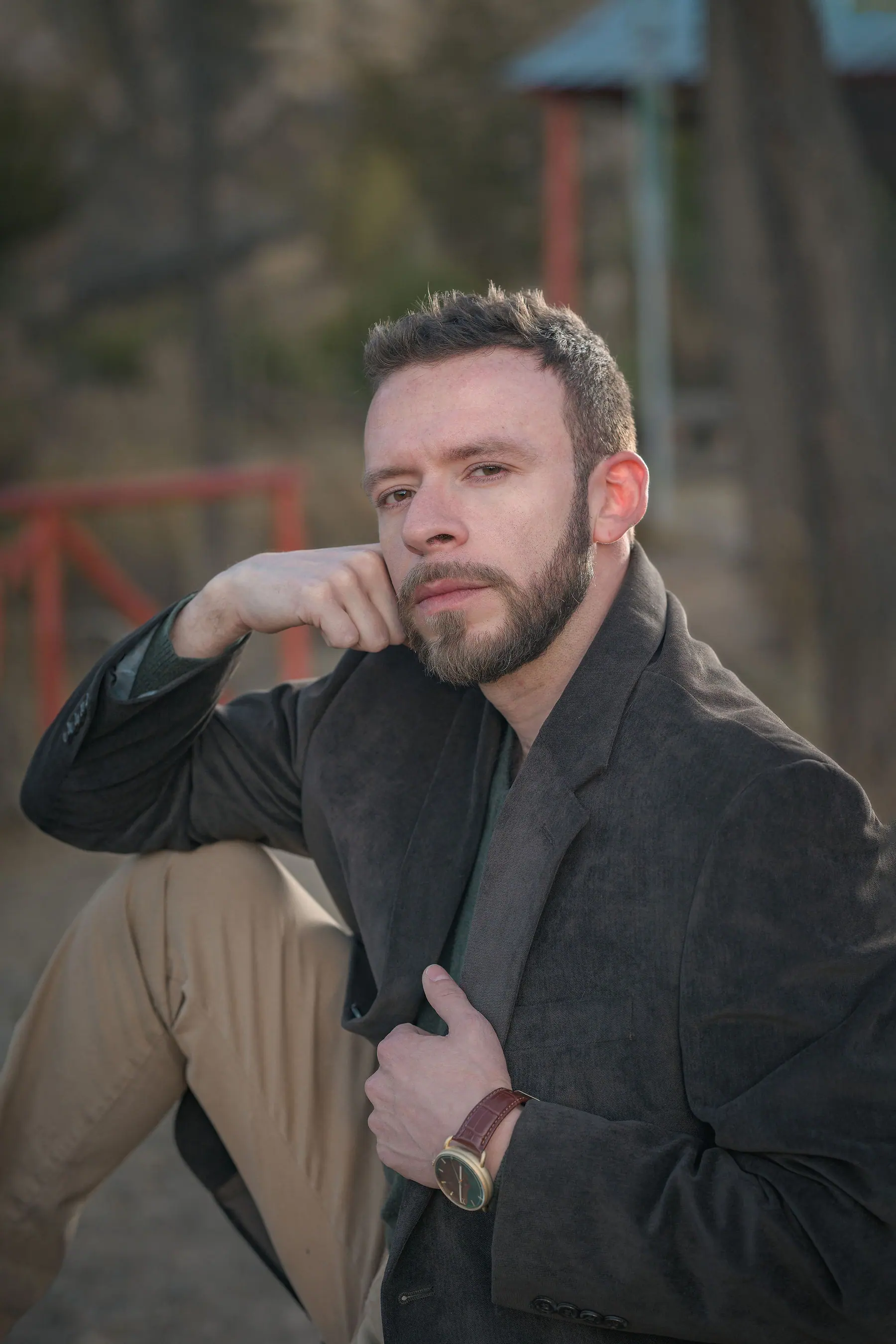Outdoors in a dark grey blazer with one leg raised, before a blurred red metal structure.
